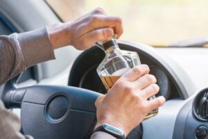 A faceless driver opens an unlabeled whiskey bottle while at the wheel of a car.